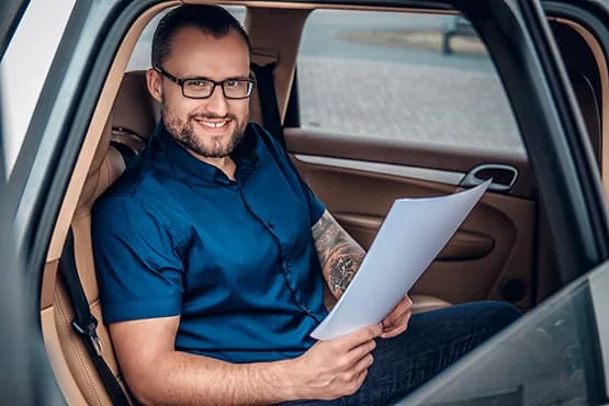 Un homme portant des lunettes est assis à l’arrière d’une voiture, des documents à la main.