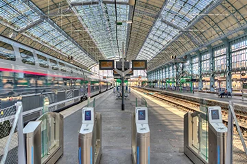 Quais et portillons d’une gare sous une grande verrière, avec un train à gauche.