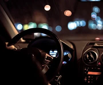 Un conducteur tient le volant d’une voiture, face aux lumières de la rue la nuit.